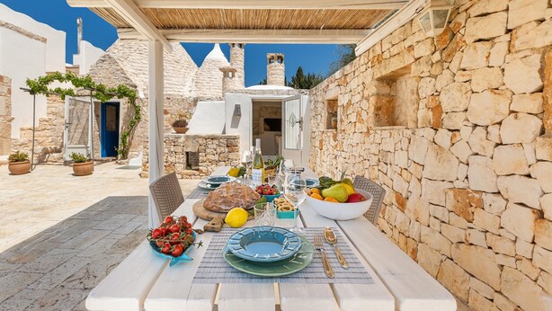 A table set for a meal with a stone wall backdrop, a thatched roof, and a view of white buildings with towers. - 2530 Trullolive by Perle di Puglia (Ostuni)