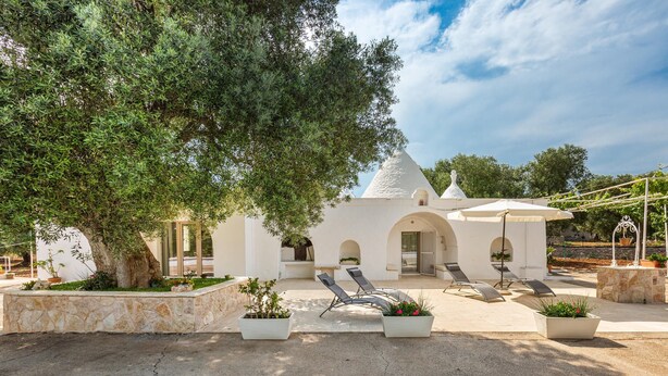 A modern villa with a white exterior, a conical roof, and a patio area with two lounge chairs and potted plants. - 2985 Trullo tra gli Ulivi by Barbarhouse (Carovigno)