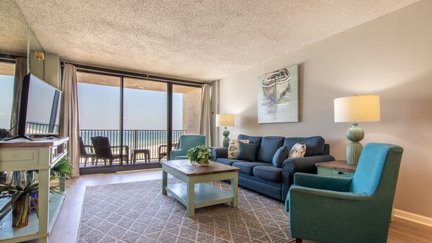 A living room with a sofa, armchair, coffee table, and a TV. There is a balcony with a view.
