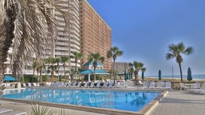 A hotel pool area with lounge chairs and umbrellas, a large building in the background, and a clear sky.