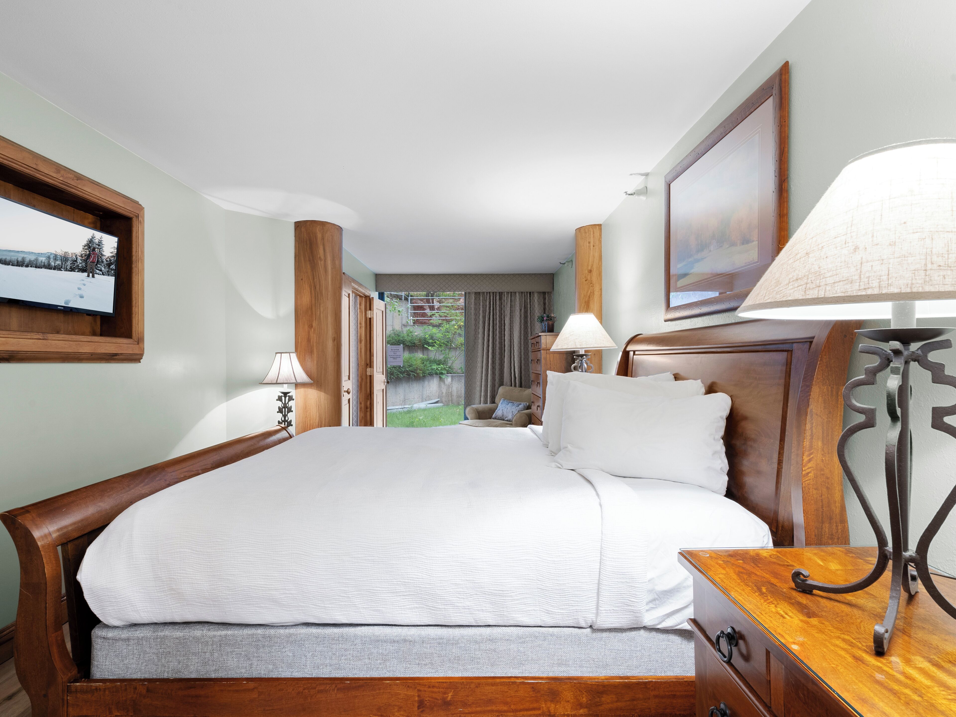 Habitación de hotel con una cama grande, cabecera de madera, dos mesitas de noche con lámparas, un escritorio pequeño y vistas a un jardín a través de una puerta abierta.