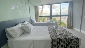 A hotel room with a large bed, a smaller bed, and a sofa. There is a window with a view of palm trees and buildings. - POUSADA AMADA TERRA (Ipojuca)