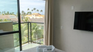 A hotel room with a large bed, a TV mounted on the wall, and a balcony with a view of palm trees and buildings. - POUSADA AMADA TERRA (Ipojuca)