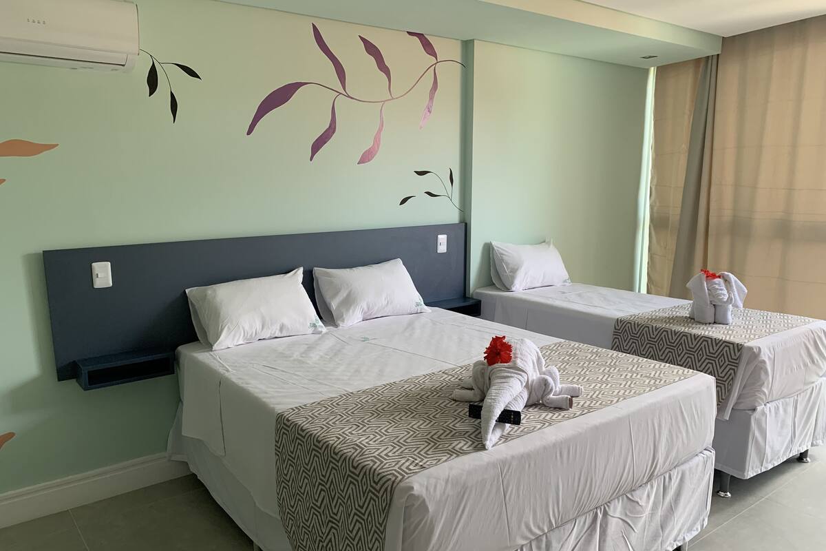 Habitación de hotel con dos camas, cada una con una colcha estampada y almohadas blancas. Decoración de hojas en la pared y una ventana con cortinas.