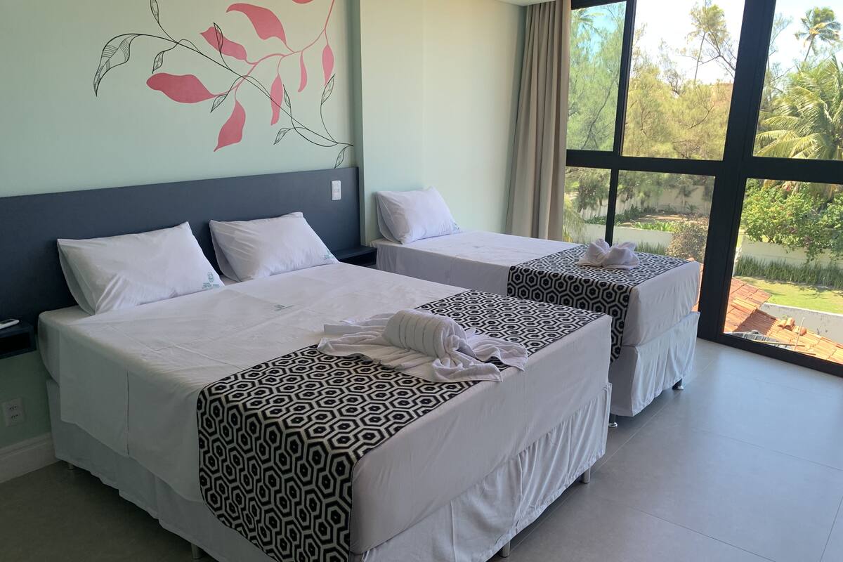 Habitación de hotel con dos camas, cada una con una colcha estampada y almohadas blancas. Un ventanal grande ofrece vistas a la vegetación exterior.