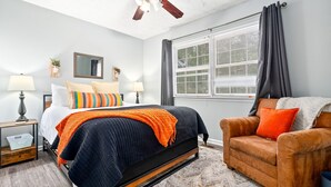 Un dormitorio con cama, mesita de noche, silla, ventana con cortinas y ventilador de techo.