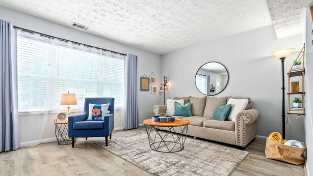 Una sala de estar con un sillón azul, un sofá beige, una mesa de centro redonda y una alfombra estampada.