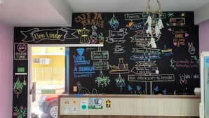 Una cafetería con una pared de pizarra que muestra diversos mensajes y dibujos, un mostrador de recepción y un banco de madera en primer plano.