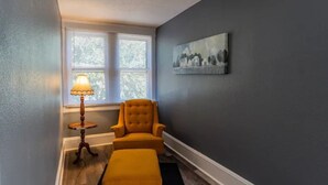 A room with a dark gray wall, a window with blinds, a yellow armchair, a small table with a lamp, and a painting.