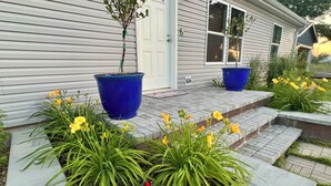 Une maison avec un porche à l’avant, une porte et une fenêtre. Des plantes et des fleurs en pot sont disposées sur le porche.
