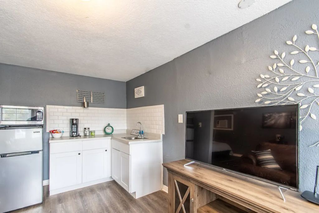 A compact kitchen with white cabinets, a microwave, a coffee maker, and a sink. There's a wooden bench and a flat-screen TV mounted on the wall.