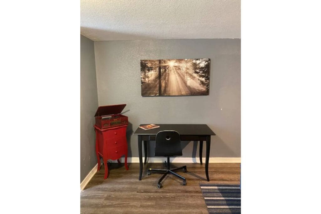 Una habitación con escritorio, silla, cómoda roja y un cuadro de un paisaje con carretera.