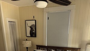 Una cama bien tendida con una colcha blanca y azul, dos almohadas oscuras y una manta azul. Se observa un cabecero de madera, una mesita de noche con una lámpara y un cuadro enmarcado en la pared.