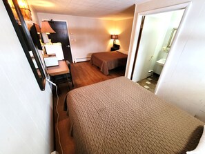 Habitación de hotel pequeña con una cama individual, un escritorio con silla, una mesita, una lámpara y un baño visible a través de una puerta abierta.