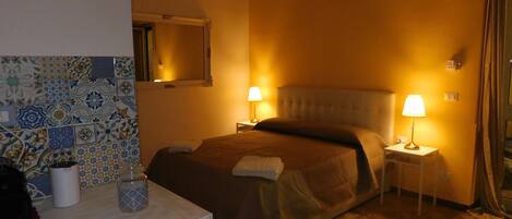 A hotel room with a bed, two bedside lamps, a nightstand, a mirror, and a tiled wall.