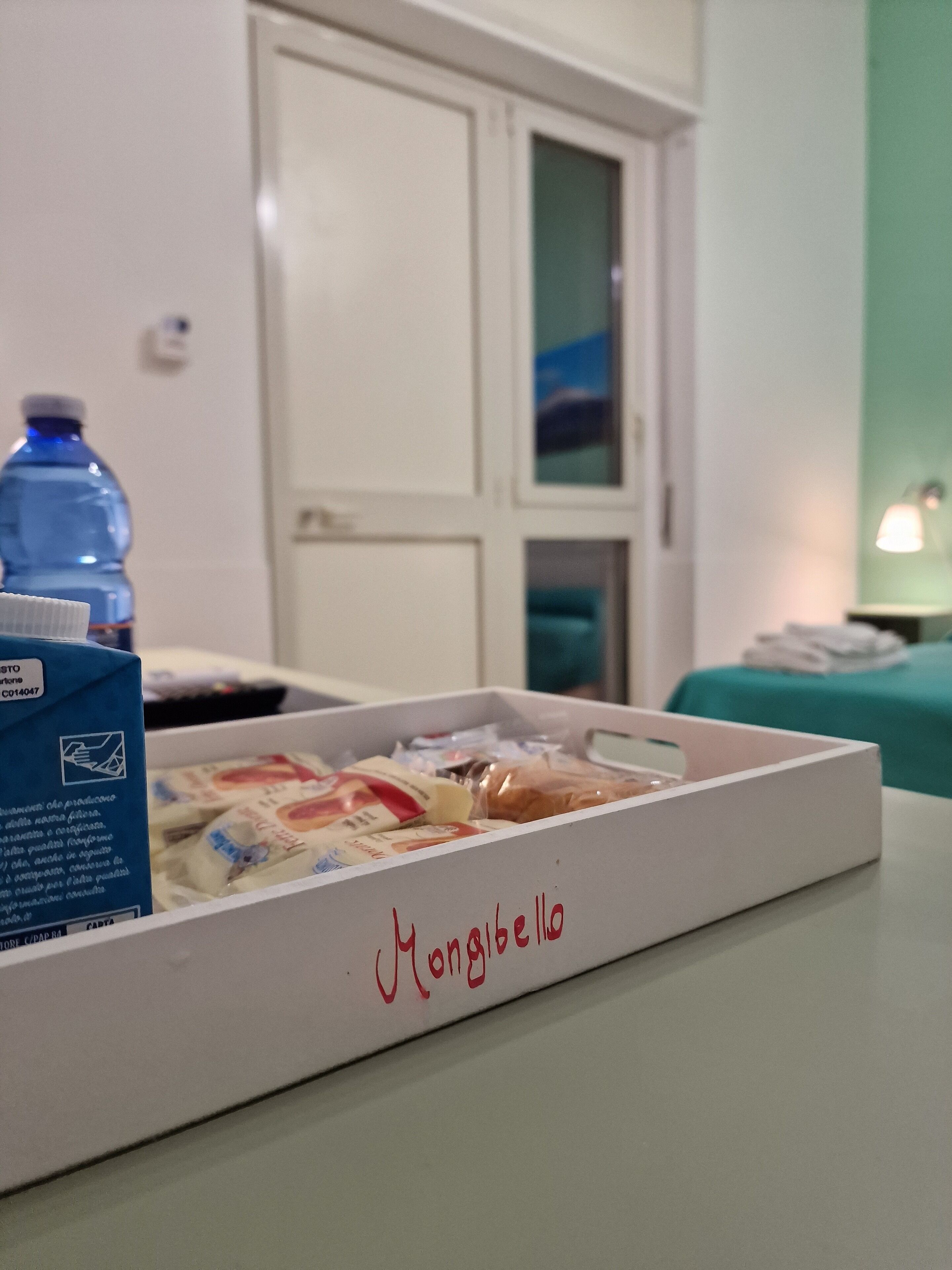 Une pièce avec un lit, une table sur laquelle se trouve une boîte de nourriture, une bouteille d’eau et une porte.