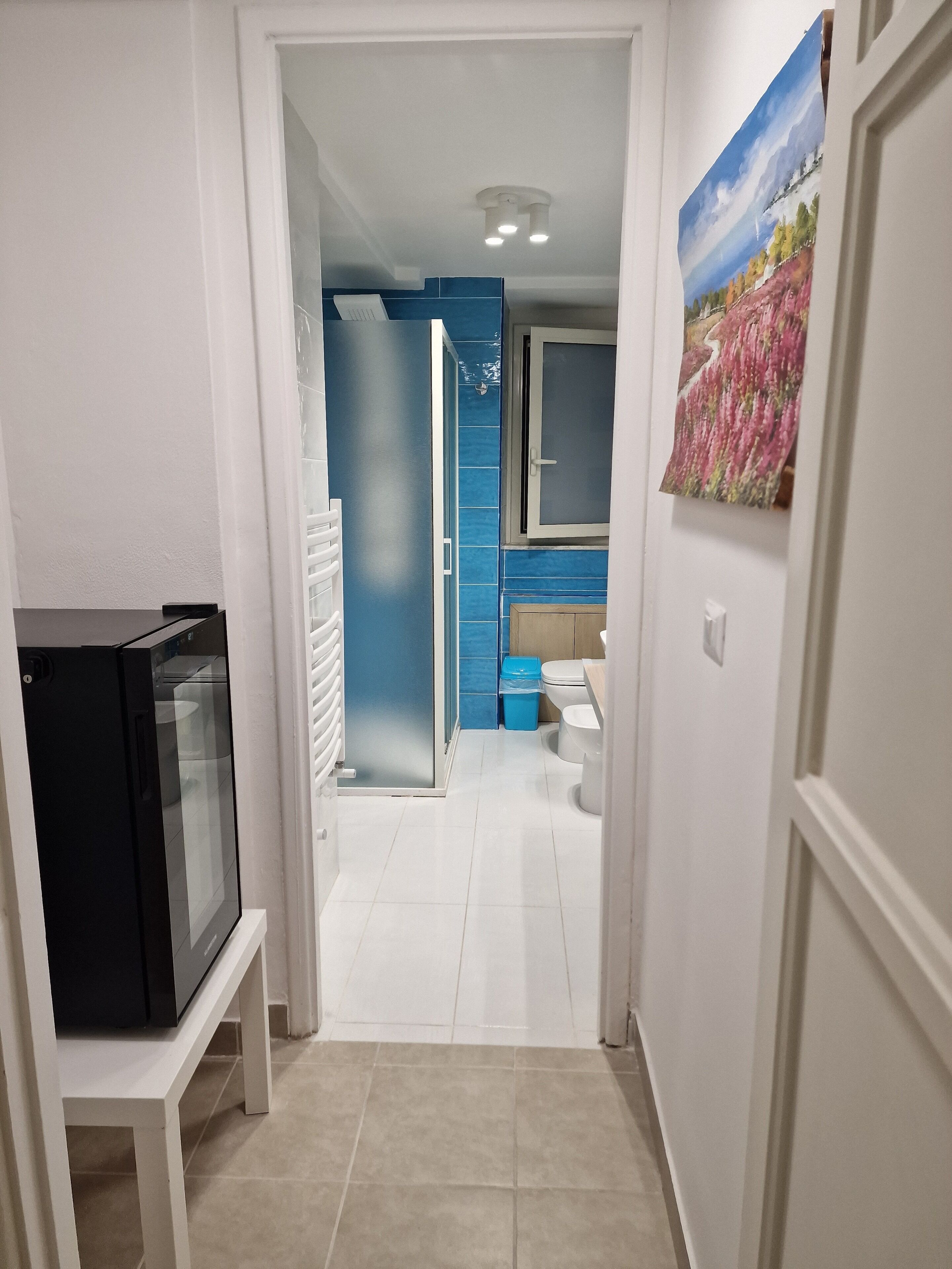 A hallway leading to a bathroom with a blue accent wall, a toilet, and a sink. There is a black refrigerator on a white stand to the left.