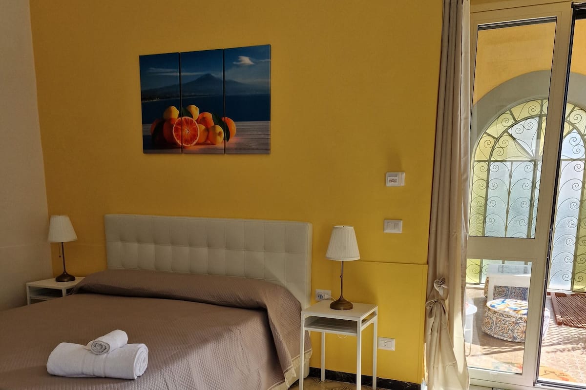 Une chambre d’hôtel avec un lit, une table de chevet, une lampe, un tableau au mur et une vue à travers une porte vitrée.