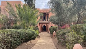 A pathway leading to a building with arches and a tiled roof, surrounded by lush greenery and a well-maintained garden. - In the heart of Marrakech's Palmeraie (Marrakesh)