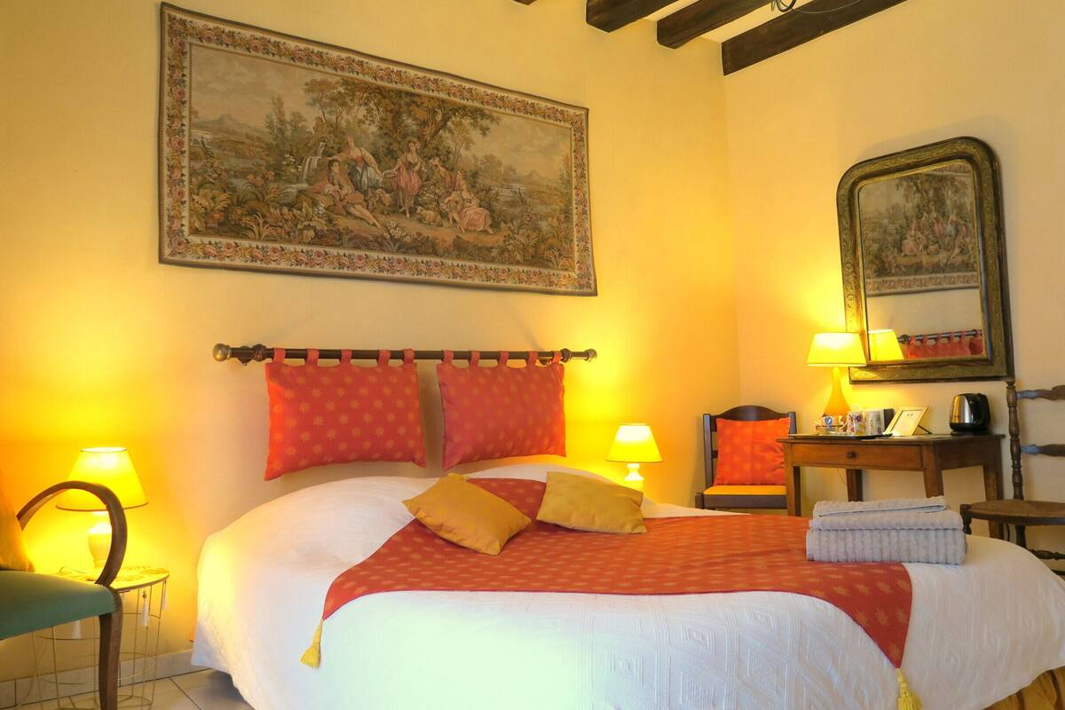 A hotel room with a large bed, two bedside lamps, a wooden chair, a small table, and a painting on the wall.