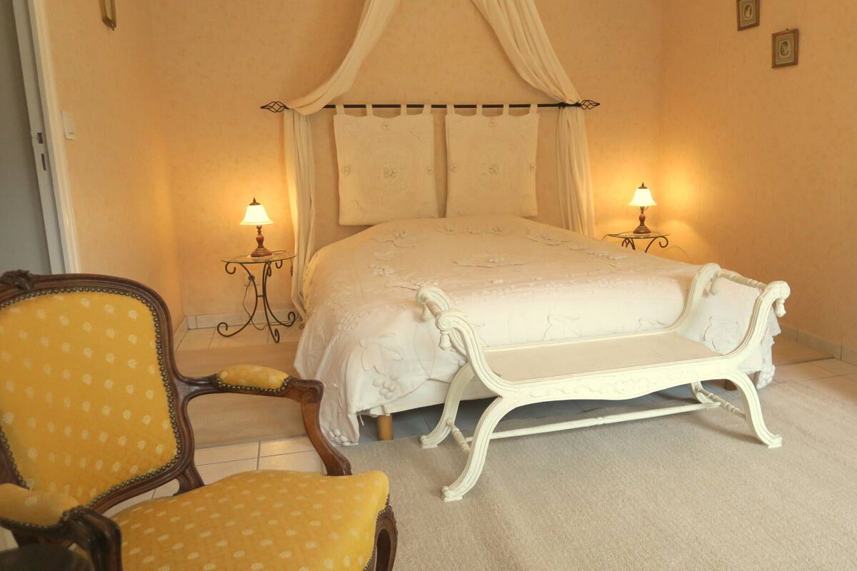 A bedroom with a four-poster bed, two bedside lamps, a wooden chair with a yellow cushion, and a white bench.