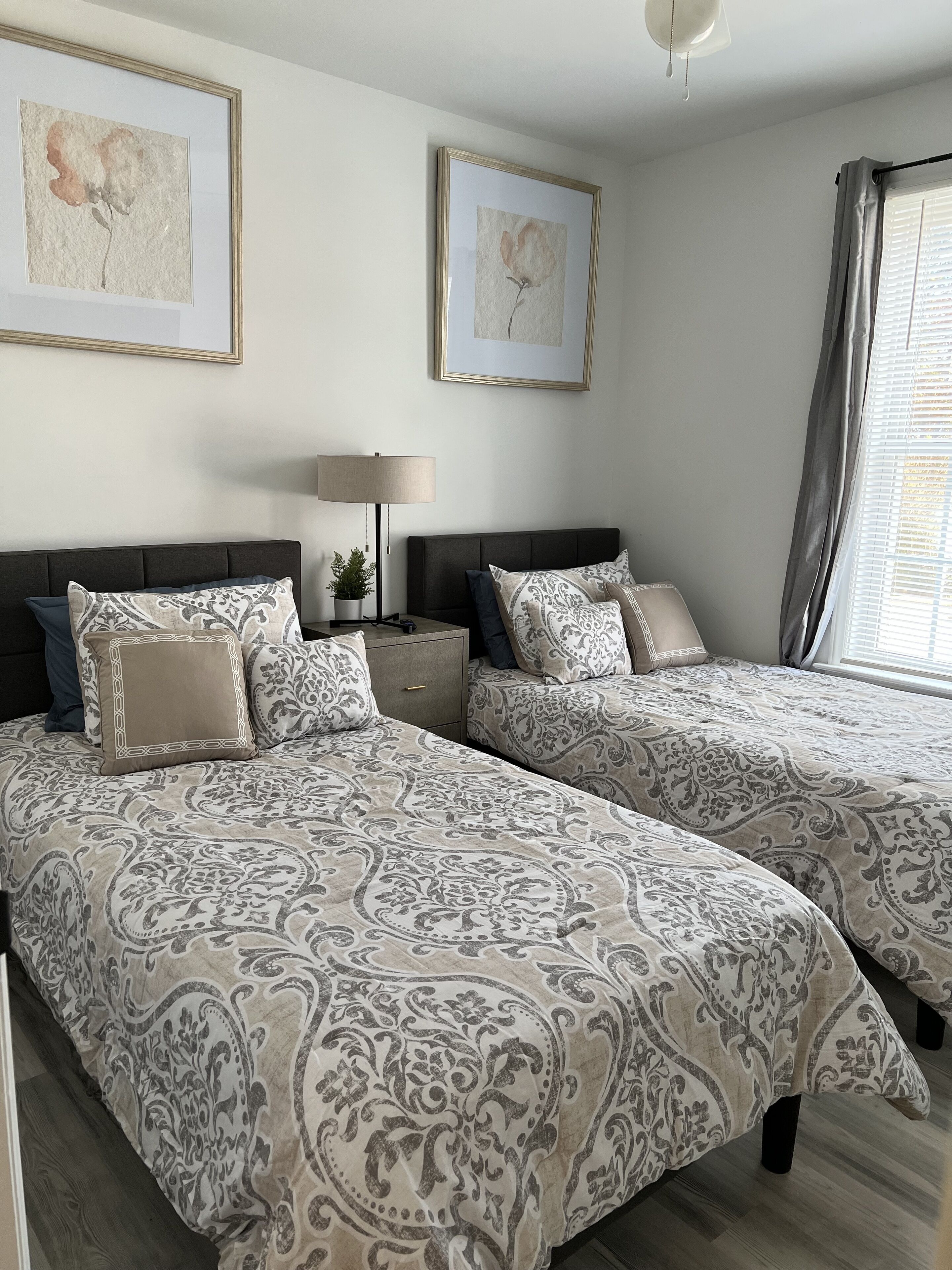 A bedroom with two beds, a bedside table with a lamp, and two framed artworks on the wall.