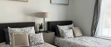 A bedroom with two beds, a bedside table with a lamp, and two framed artworks on the wall.