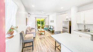 Una sala de estar moderna con sofá, mesa de centro y televisor. Se observa una cocina con refrigerador, microondas y pileta. Se ve un balcón con plantas a través de la ventana.