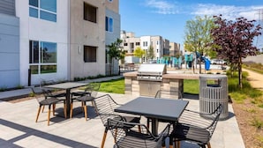 En udendørs terrasse med grill, siddepladser og en legeplads i baggrunden.