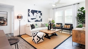 En moderne stue med en hvid sofa, en skammel med sort og hvidt mønster, et træbord og en sort sofa med puder. Der er et stort abstrakt maleri på væggen, en gulvlampe og en potteplante.