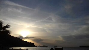 Escena de platja amb el sol ponent sobre l'aigua, siluetes de palmeres i formes de terra a la distància.
