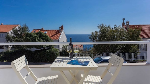 A balcony with a white table and chairs, overlooking a coastal view with red-roofed buildings and the sea.