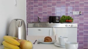 A kitchenette with a toaster, kettle, and a small stove. There are fruits, a croissant, and a cup on the counter.