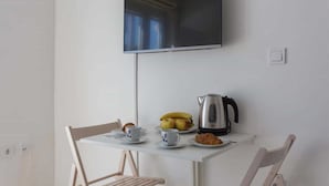 A small dining area with a white table, two chairs, a television mounted on the wall, a kettle, a plate with bananas and apples, a cup, and a croissant.