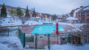 Une piscine entourée d’une zone enneigée, avec une clôture, un parasol rouge et un bâtiment en arrière-plan.