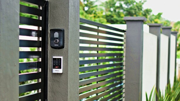 A modern gate with a keypad and a digital display, flanked by vertical columns and a metal fence with horizontal slats. - MORADA PINHEIRO (Bombinhas)