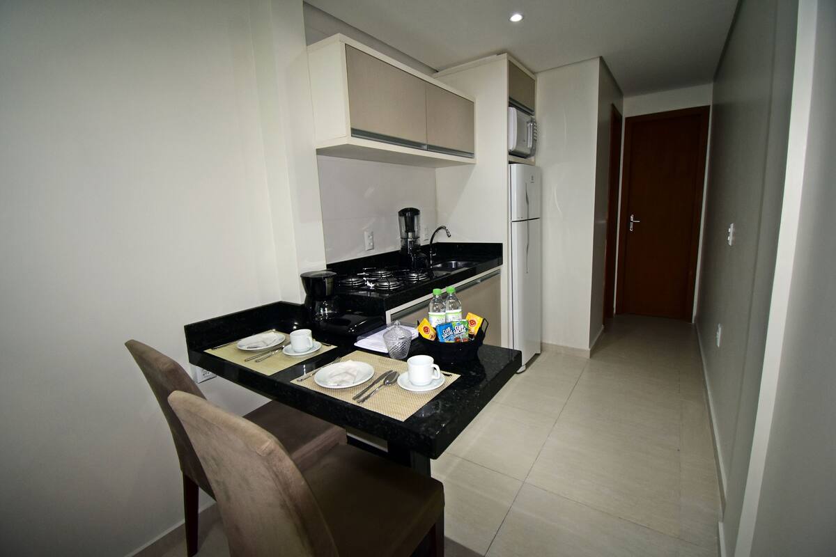 Une cuisine compacte avec un plan de travail noir, une cuisinière et un réfrigérateur. On y trouve une table à manger avec des chaises et un petit meuble de rangement au-dessus.