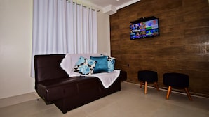 A living room with a dark brown sofa, two black ottomans, a flat-screen TV on a wooden wall, and a white curtain. - MORADA PINHEIRO (Bombinhas)