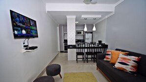 A modern living room with a flat-screen TV, a dark sofa with patterned cushions, a small rug, and a dining area with chairs and a table. - MORADA PINHEIRO (Bombinhas)