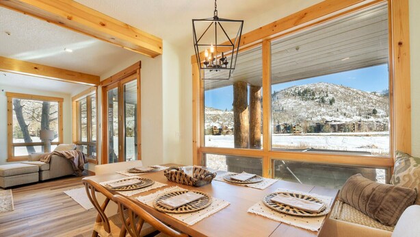 A cozy dining area with a wooden table set for four, large windows offering a view of a snowy landscape, and a rustic chandelier hanging above.