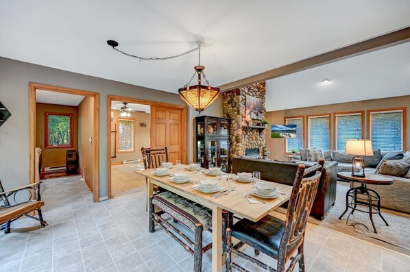 A spacious dining area with a long table set for four, surrounded by wooden chairs. A stone wall and a fireplace are visible in the background.