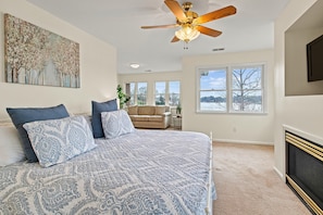 A bedroom with a large bed, a ceiling fan, a fireplace, and a view of the outdoors.