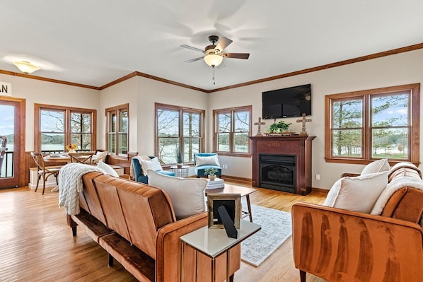 A spacious living room with a fireplace, large windows, and a ceiling fan.