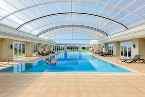 An indoor swimming pool with a glass ceiling, clear views of the outdoors, and sun loungers around the pool. - Boavista Golf and Spa Resort - Bayview (Lagos)