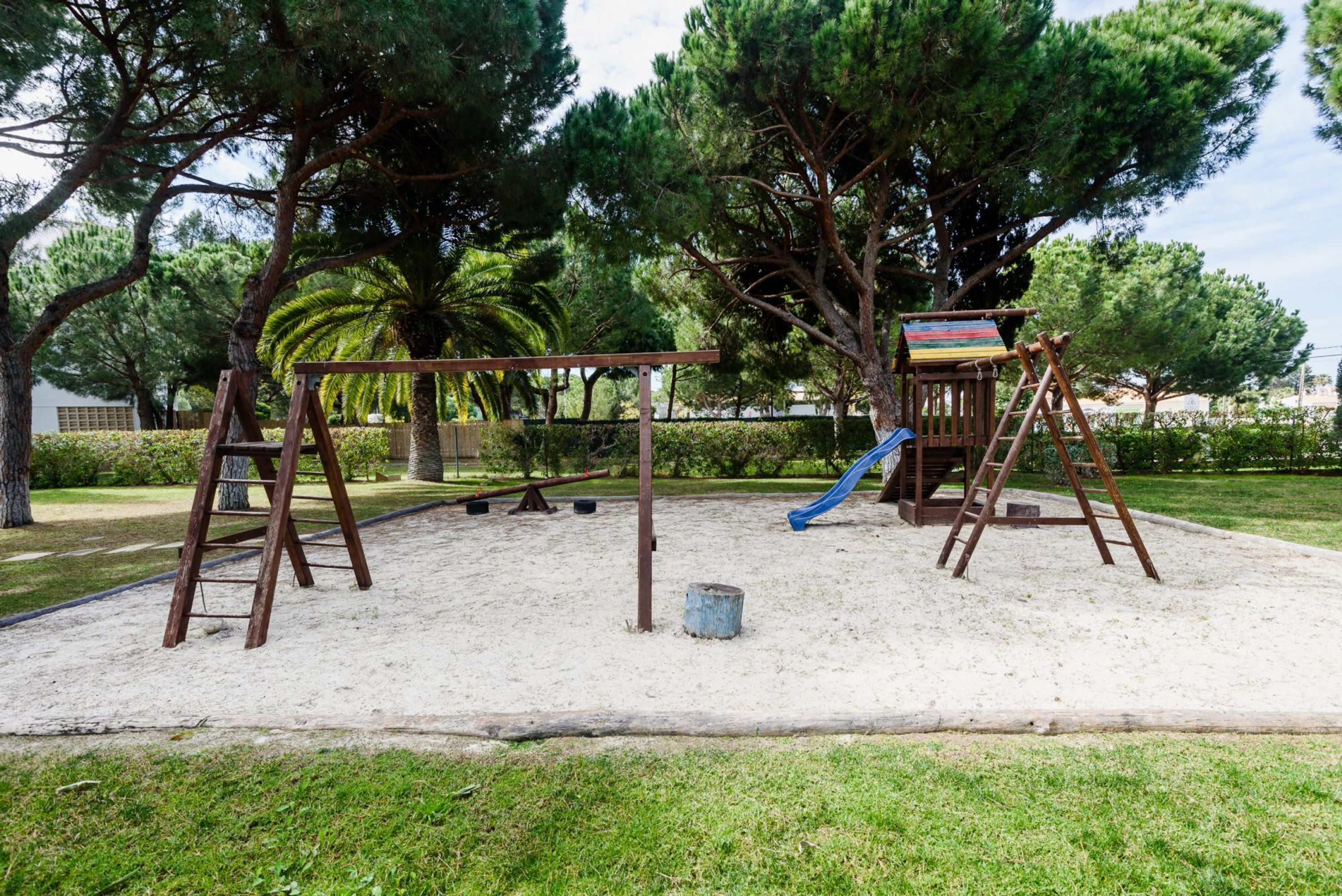 Zona de jocs amb cadira de balanç, tobogan i estructura per escalar en un parc amb arbres i sorra.