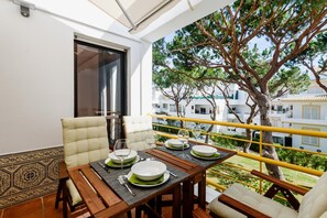 A balcony with a dining table set for four, surrounded by white cushioned chairs, overlooking a tree and a building. - Aldeia das A Oteias Apartment (Albufeira)