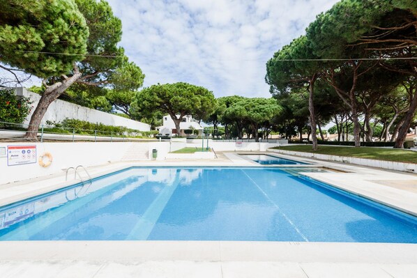 A swimming pool with clear blue water, surrounded by trees and a white building in the background. - Aldeia das A Oteias Apartment (Albufeira)