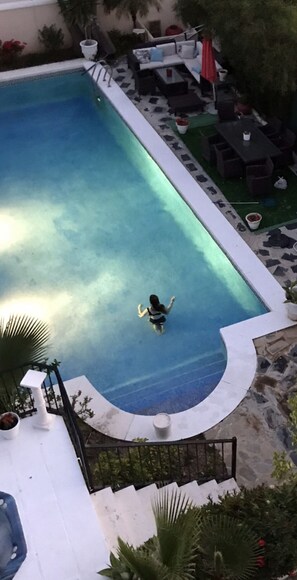 Une piscine avec une personne dans l’eau, entourée de chaises longues et d’une petite zone aménagée pour se détendre.