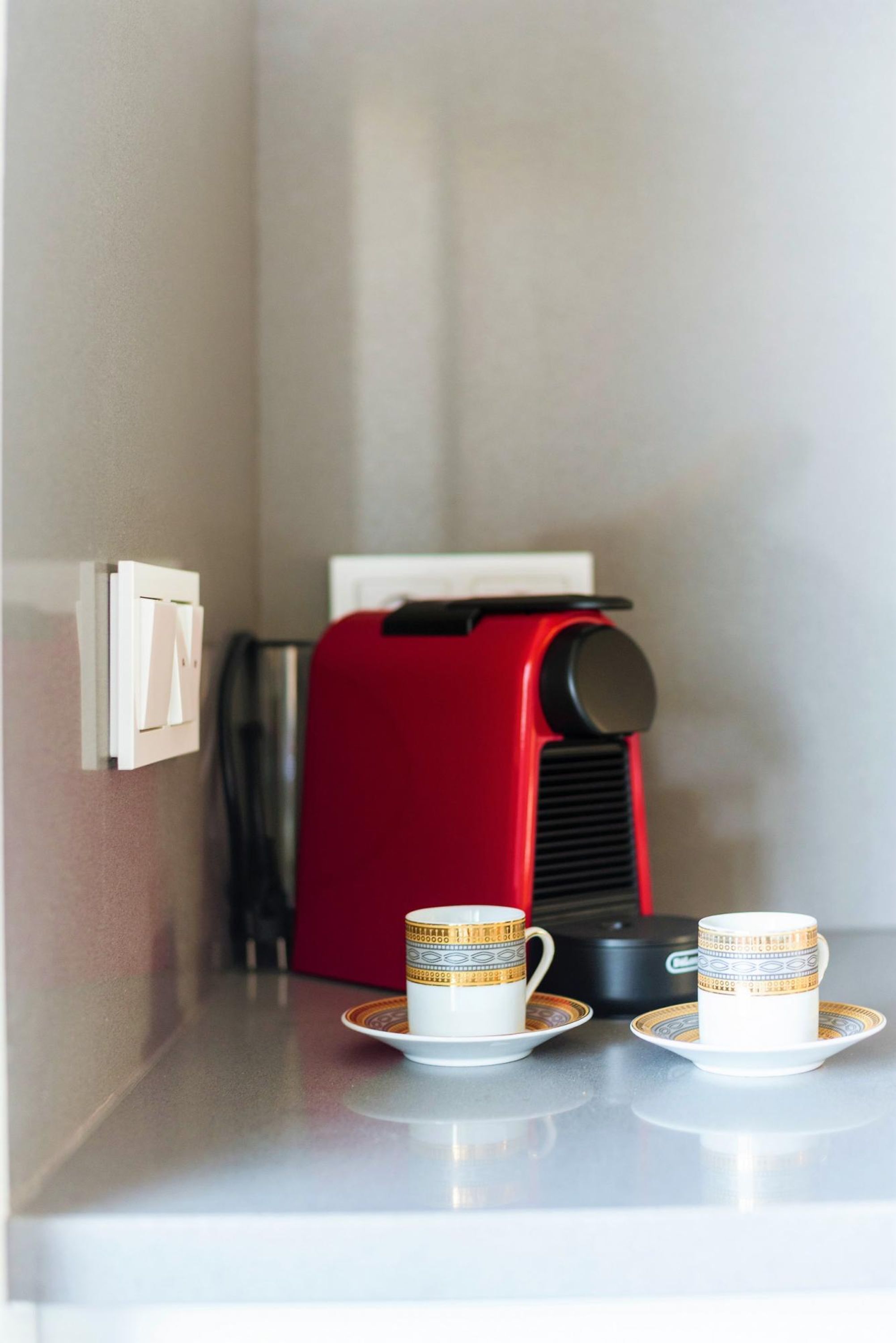 Een kleine keuken met een rode koffiezetapparaat, twee kopjes op schoteltjes en een lichtschakelaar aan de muur.