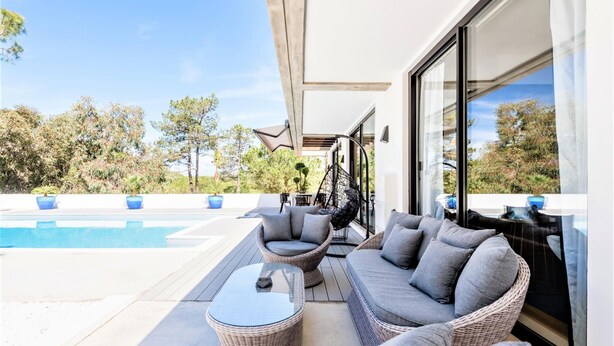 Uma área de estar moderna ao ar livre, com piscina, móveis de vime e vista para árvores e céu.